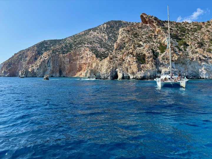 A sailboat moored beneath tall sunlit cliffs in deep blue water.